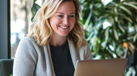 Een vrouw in de dertig met blond haar en een grijze vest en zwart shirt die glimlachend op een macbook werkt.